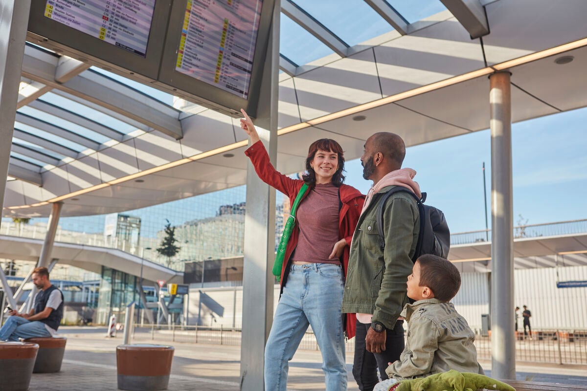 Een gezin staat op het perron en kijkt naar een digitaal reisinformatie bord